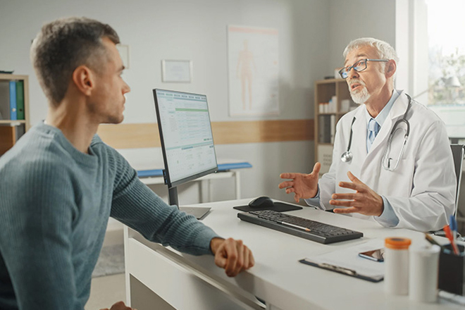 Healthcare professional consulting with a man during an appointment, with a computer screen visible.