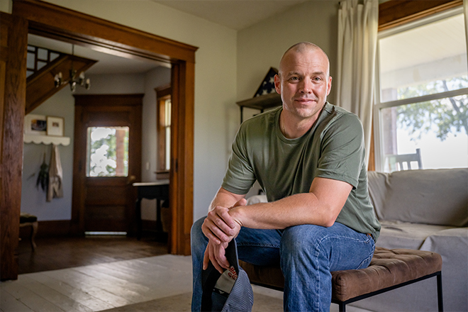 Casually dressed man in a green shirt posing in a living room with natural light.
