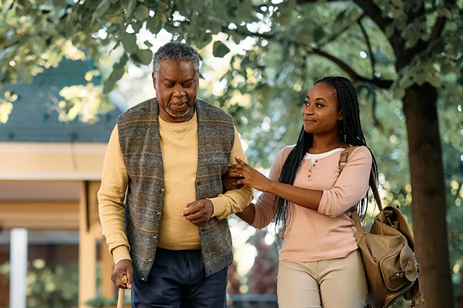 Senior man walking with support from a younger woman outdoors under trees.