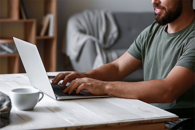 Man smiling while typing on a laptop at a home desk with a coffee cup.