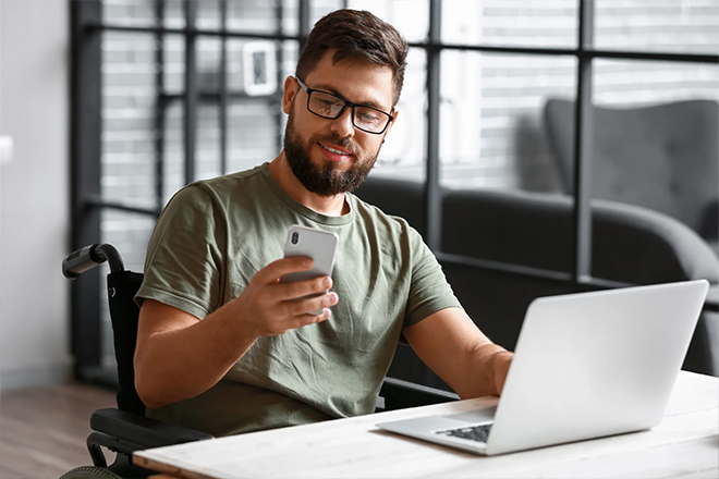 Smiling man in glasses sitting at a desk, checking his phone while working on a laptop.