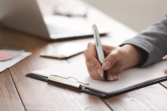 Person filling out paperwork at a desk with a laptop in the background.