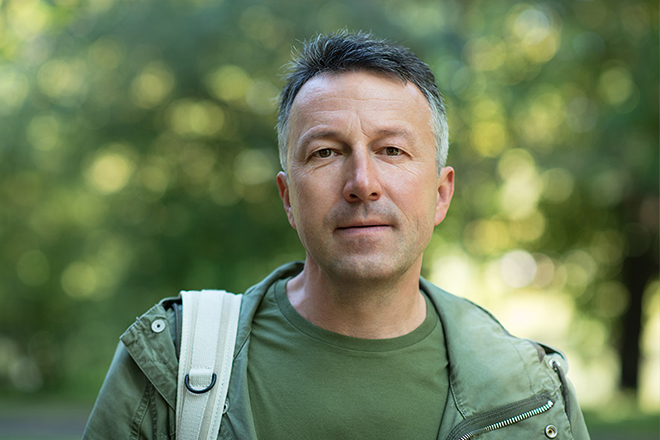 Middle-aged man outdoors with a neutral expression, wearing a green jacket and backpack.